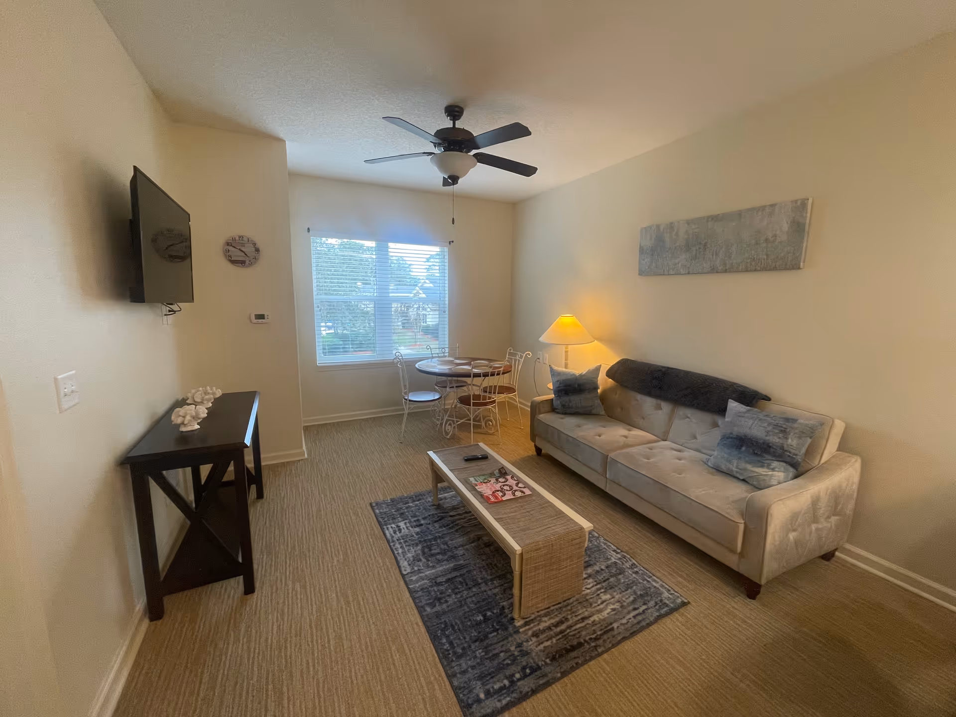 A cozy living room with a beige sofa adorned with two blue pillows and a dark throw blanket. A small wooden coffee table with magazines and a remote control sits on a patterned rug in front of the sofa. To the left, there is a black console table with decorative items and a wall-mounted flat-screen TV above it. A round dining table with four chairs is positioned near a window with white blinds, allowing natural light to brighten the room. A ceiling fan with a light fixture hangs from the ceiling, and a table lamp on a side table next to the sofa provides additional warm lighting. The walls are painted a light beige color, and a rectangular piece of abstract art hangs above the sofa.