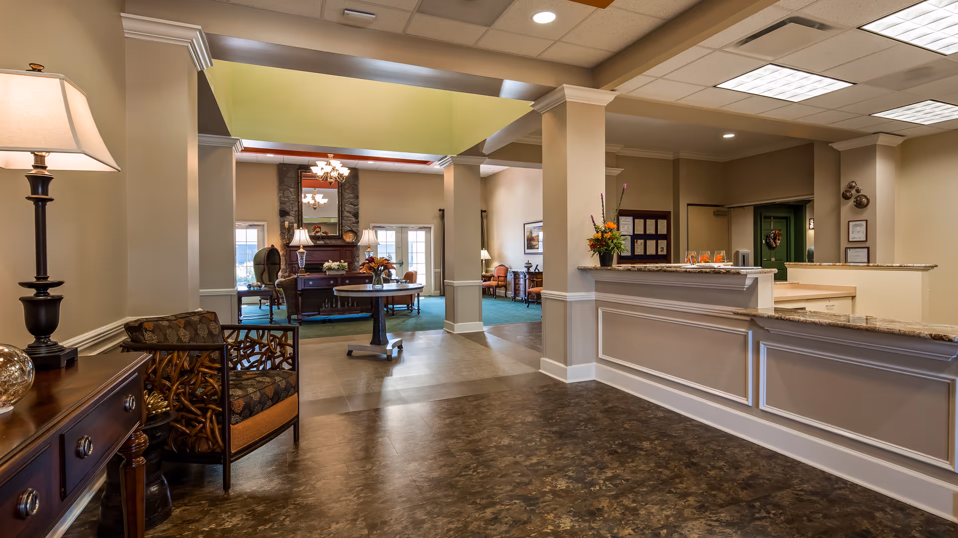 Interior view of a senior living facility lobby area with a reception desk on the right, a seating area with chairs and tables in the background, warm lighting, and decorative elements including flowers and lamps.