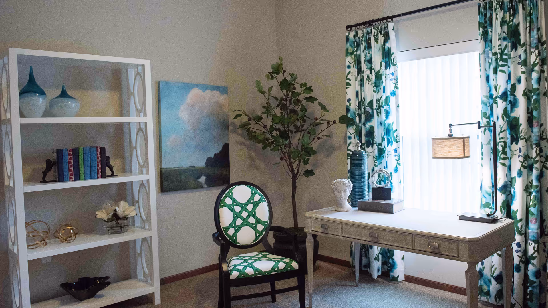 Bright furnished sitting area with a desk, decorative chair, white bookshelf, potted plant, and patterned curtains by a window.