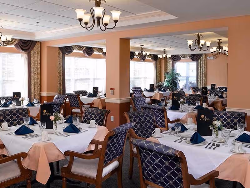 A spacious dining room with multiple tables covered in white and peach tablecloths, each set with blue folded napkins, glassware, silverware, and small floral centerpieces. The room features large windows with curtains, chandeliers hanging from the ceiling, and upholstered chairs with a blue patterned fabric.