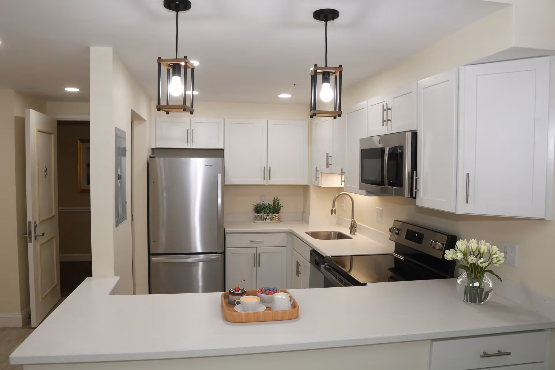 Modern kitchen with white cabinets, stainless steel refrigerator, microwave, and stove. Two pendant lights hang from the ceiling. A small tray with a cup, bowl of berries, and a dessert is on the countertop, along with a vase of white flowers.