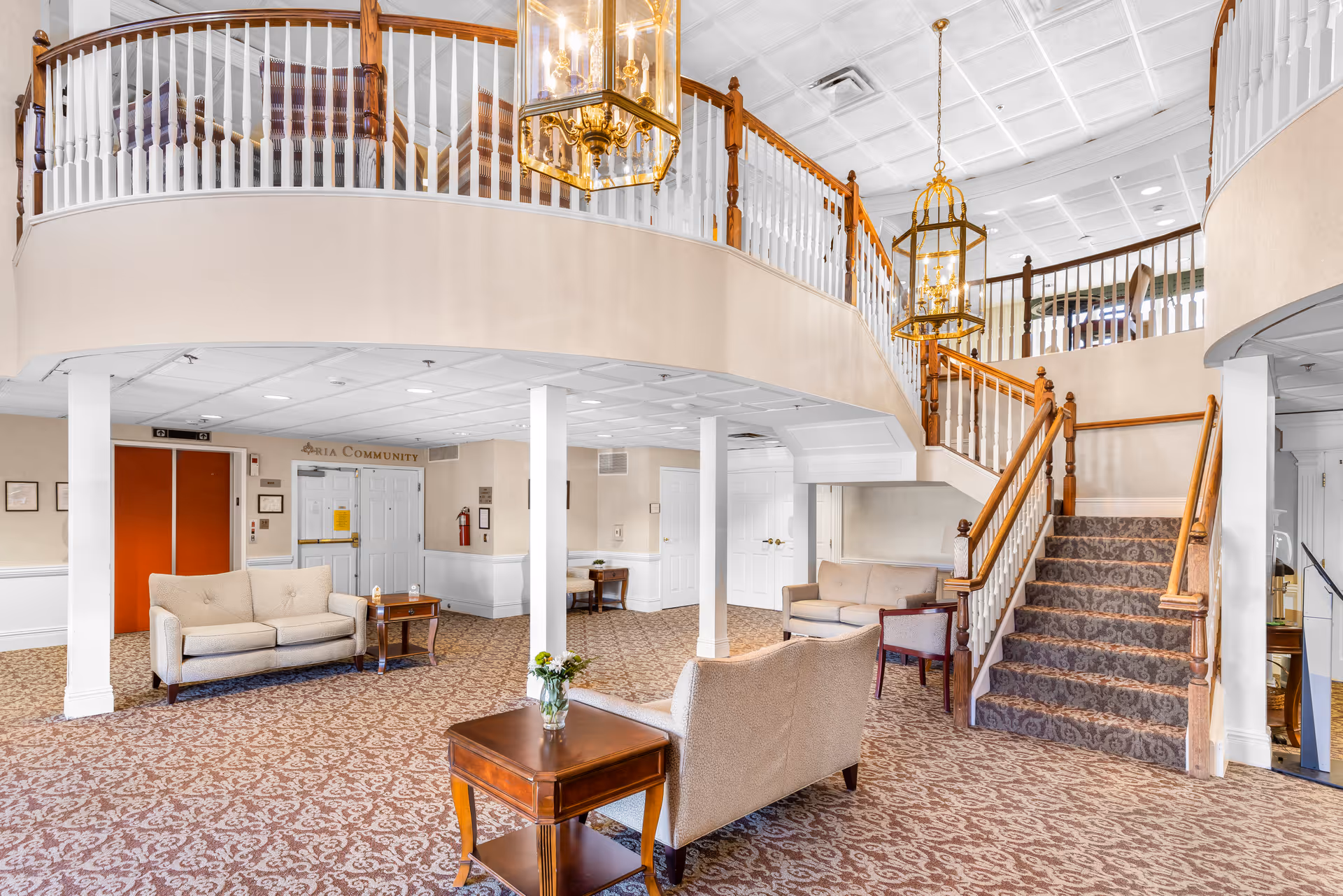 A spacious and well-lit senior living facility lobby area with patterned carpet flooring, beige walls, and white pillars. The room features multiple beige upholstered sofas and wooden tables with flower vases. A staircase with wooden handrails and carpeted steps leads to an upper balcony with white spindles and wooden railings. Two large brass and glass hanging lanterns provide lighting. There are double doors and an elevator with orange doors visible in the background.