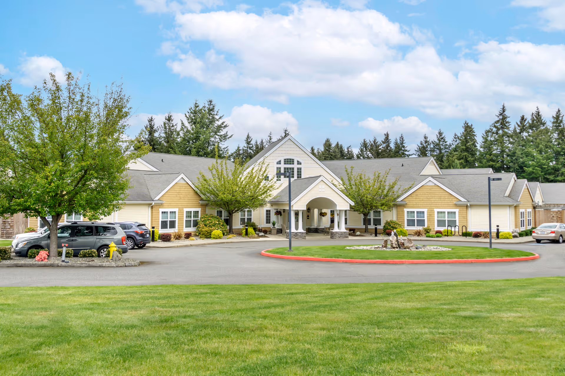 Front exterior view of a single-story senior living facility building with a covered entrance, surrounded by trees, parked cars, and a circular driveway with a landscaped island in the center. The sky is partly cloudy.