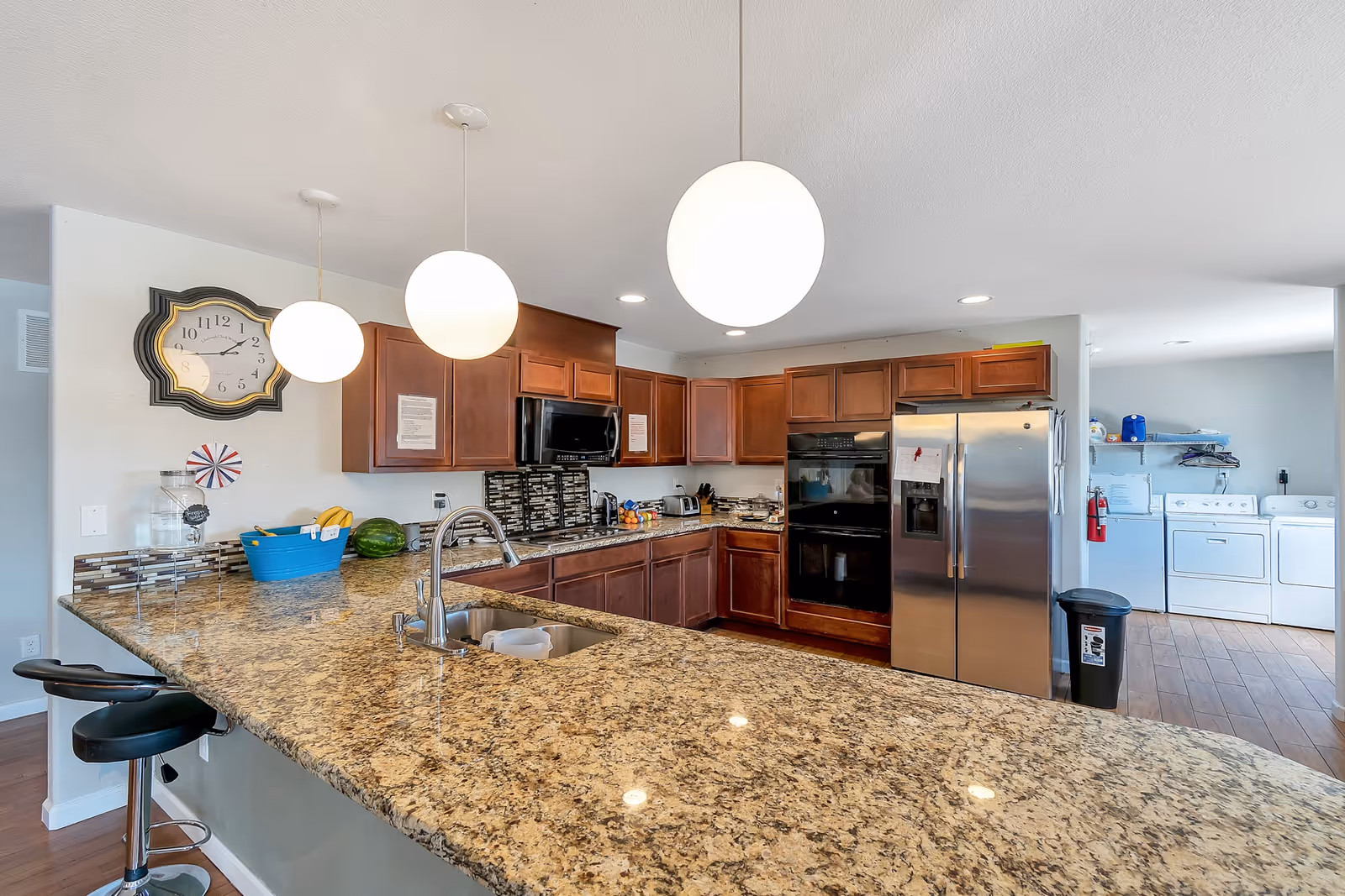 A spacious kitchen with granite countertops and dark wooden cabinets. The kitchen features a double sink, a stainless steel refrigerator, a built-in oven, a microwave, and a stovetop. Three round pendant lights hang above the countertop. In the background, a laundry area with a washer and dryer is visible. A large wall clock is mounted on the left wall, and there are two black bar stools at the counter.