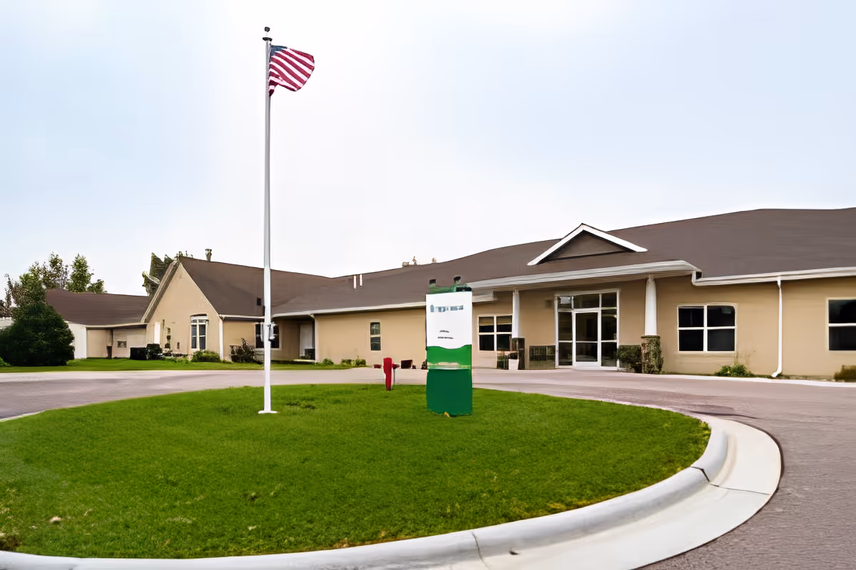 Exterior view of Edgewood Prairie Crossings Assisted Living facility showing a single-story beige building with a brown roof, a circular driveway, a green lawn in the center with a flagpole flying the American flag, and a signpost near the entrance.