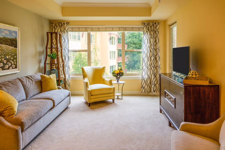 Bright living room with a sofa, yellow armchair by a large window, TV on a wooden cabinet and decorative shelving.