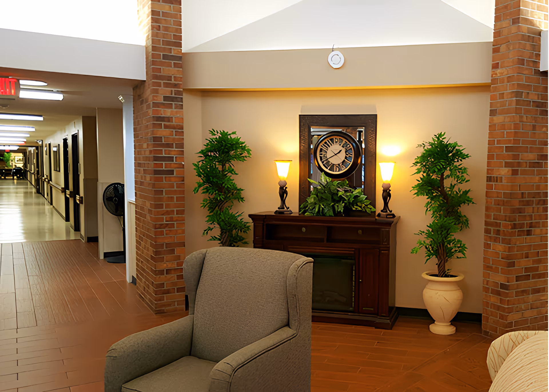 A cozy corner in a senior living facility featuring a gray upholstered armchair in the foreground, a wooden console table with a decorative clock and two lamps, flanked by two potted green plants. The area has brick pillars and a hallway with doors and handrails visible in the background.