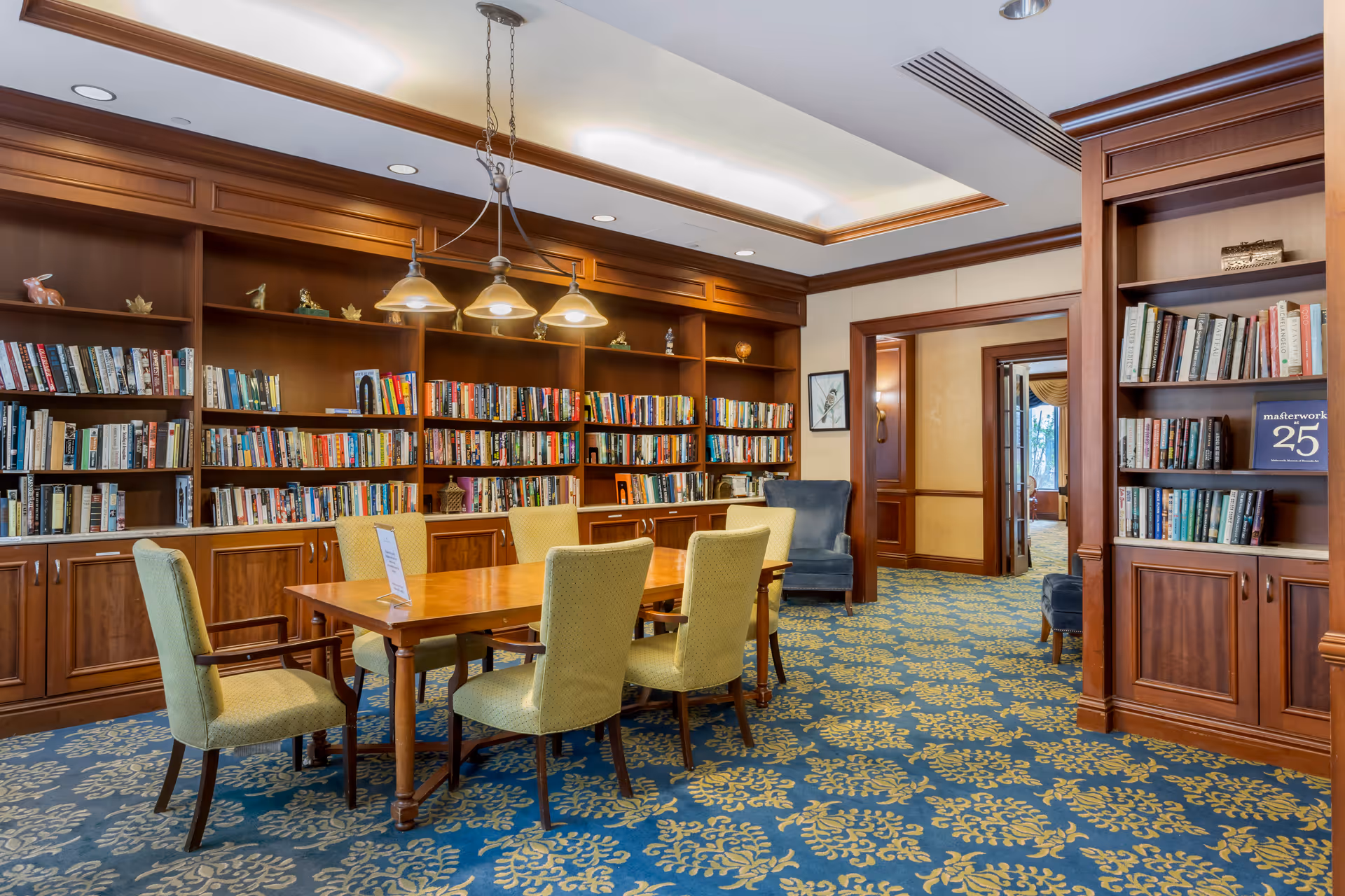 A cozy library room with wooden bookshelves filled with books along the walls. A wooden table with six upholstered chairs is centered under a hanging light fixture. The room has a patterned blue and yellow carpet and an open doorway leading to another room.