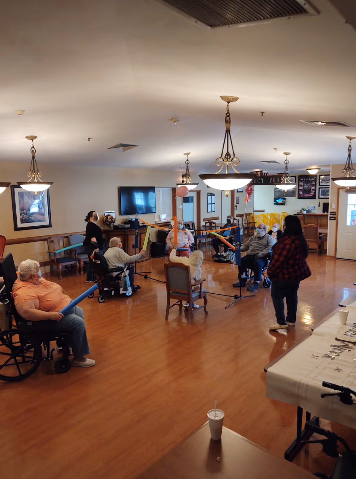 A group of elderly people, some in wheelchairs, participating in a seated balloon volleyball game in a spacious room with wooden floors and hanging light fixtures. A few staff members are present, and the room has a television, chairs, and tables with cups and utensils.