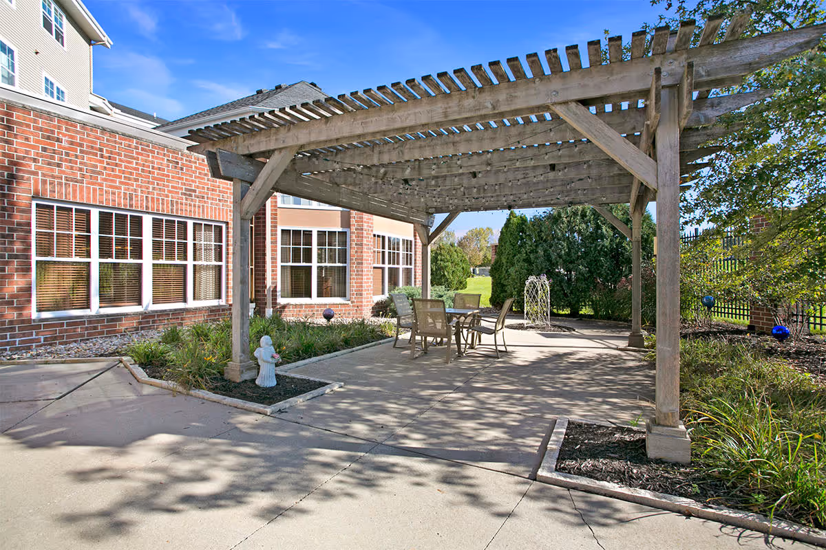 Outdoor patio area with a wooden pergola providing partial shade over a table and four chairs. The patio is surrounded by greenery, including bushes and trees, and is adjacent to a brick building with multiple windows. The sky is clear and blue.