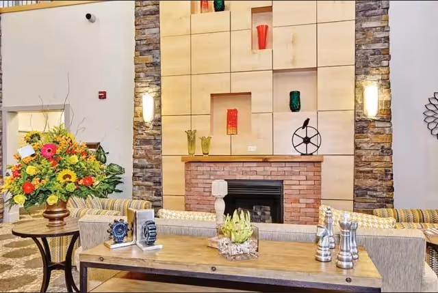 A cozy living room area featuring a brick fireplace with a wooden mantel, decorated with colorful vases and a decorative wheel. The fireplace is flanked by stone columns with wall sconces. In front of the fireplace, there is a wooden table with decorative items including a small sculpture and a glass container with plants. To the left, a round table holds a large bouquet of vibrant flowers. The seating area includes patterned sofas and cushions.