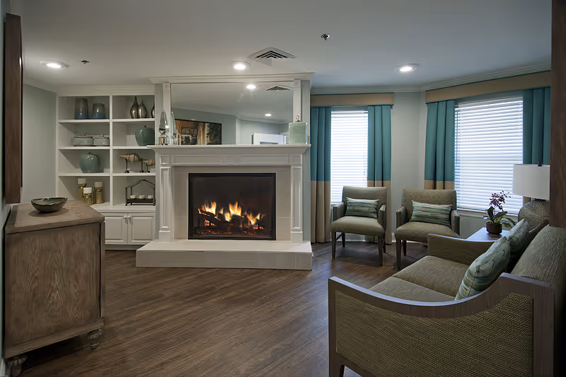 A cozy living room with a lit fireplace in the center, flanked by built-in white shelves with decorative vases and items. There are two armchairs with cushions near two windows with teal and beige curtains, and a sofa with patterned pillows. A wooden cabinet and a side table with a lamp and a small plant complete the room, which has wood flooring and recessed ceiling lights.