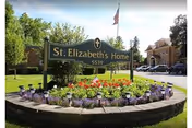 Outdoor view of St. Elizabeth's Home sign surrounded by a circular flower bed with colorful flowers and small American flags. In the background, there is a building and a parking area with cars, under a partly cloudy sky.