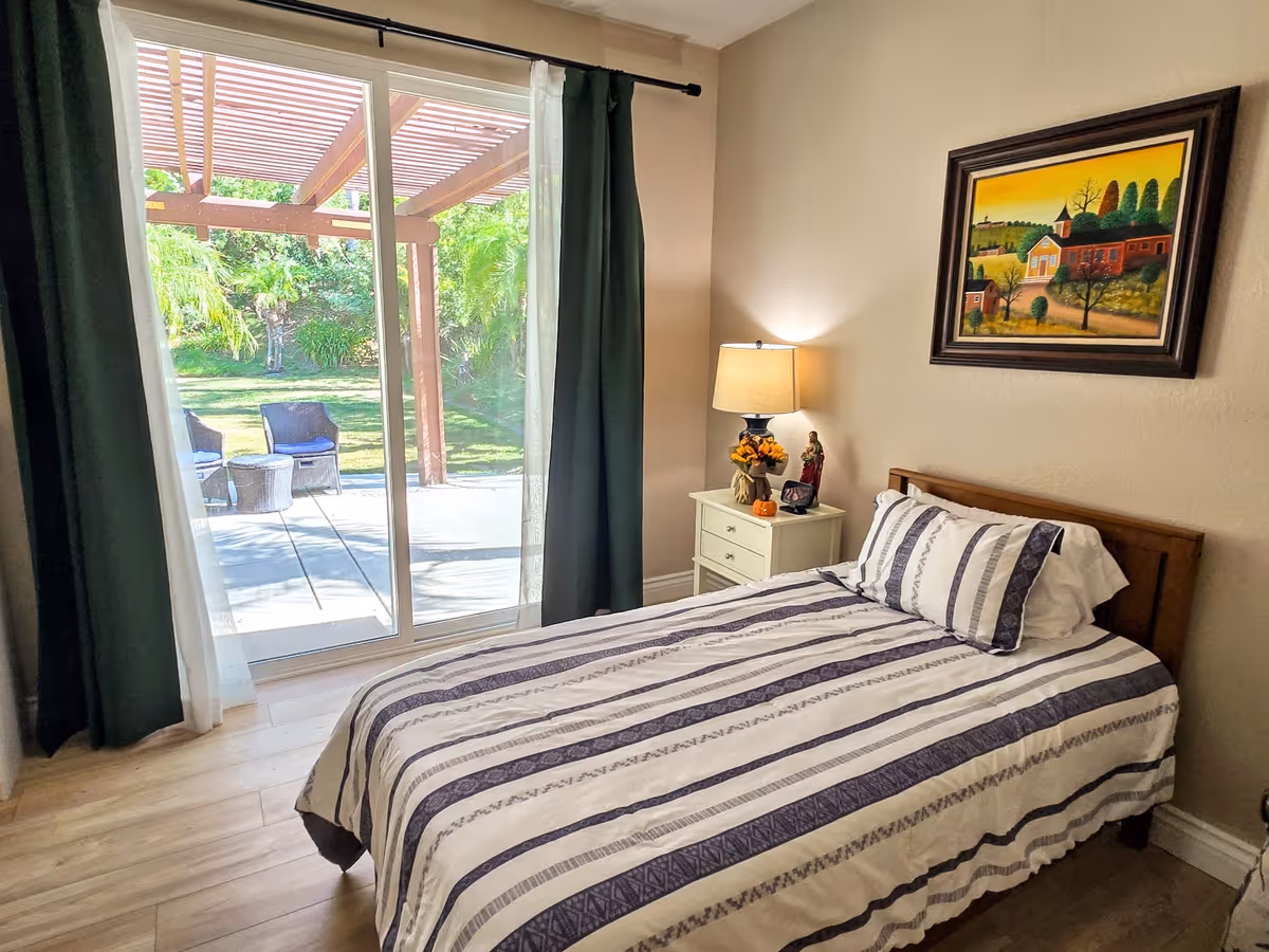 A cozy bedroom with a single bed covered in a striped bedspread. Next to the bed is a white nightstand with a lamp, a small statue, and a flower arrangement. Above the bed hangs a framed painting of a countryside scene. The room has light-colored walls and wood flooring. A large sliding glass door with dark green curtains opens to an outdoor patio with chairs and greenery.