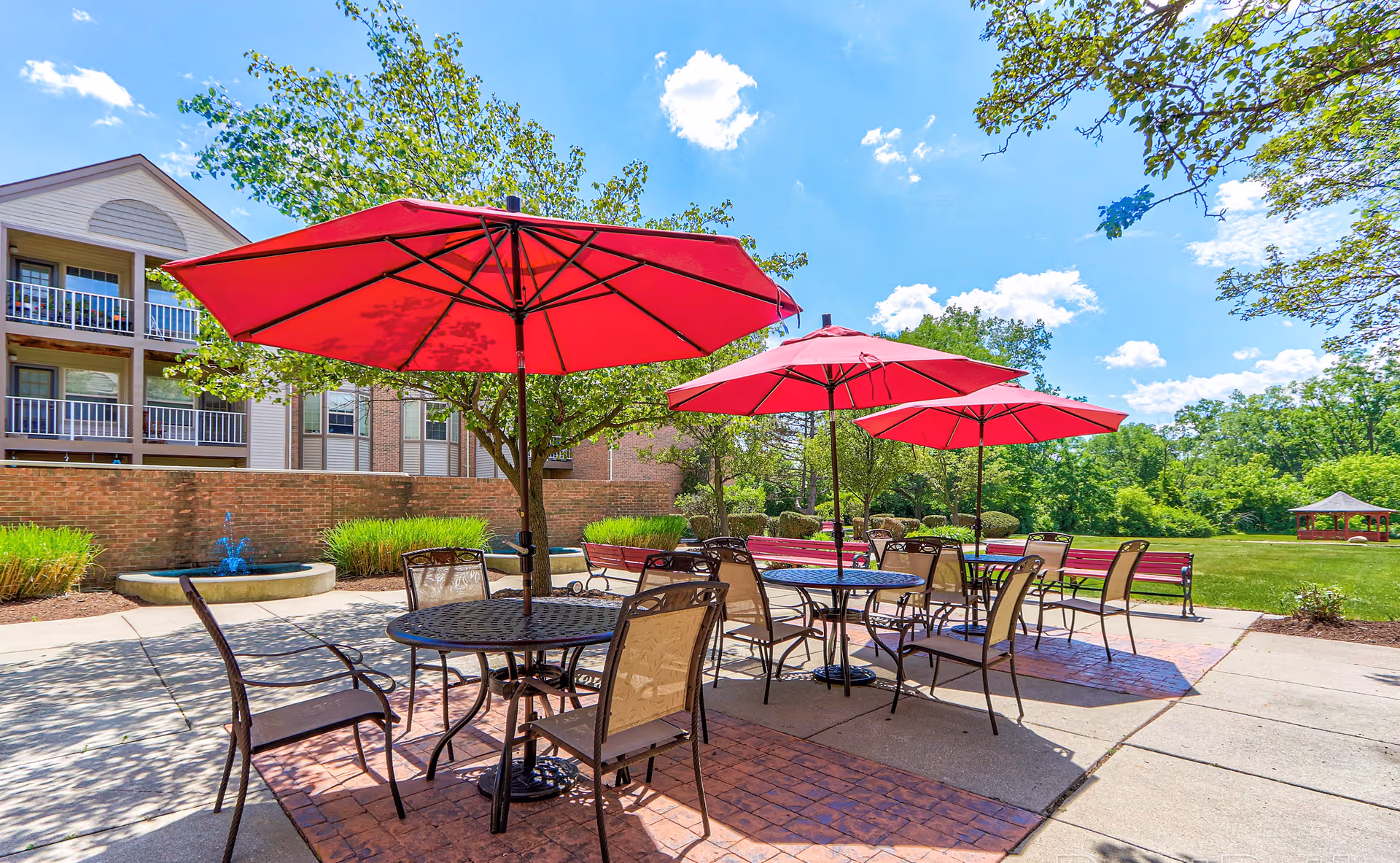 Outdoor patio area with several round tables and chairs under large red umbrellas. In the background, there is a brick building with balconies, a small water fountain, green trees, and a gazebo on a sunny day with a blue sky and scattered clouds.