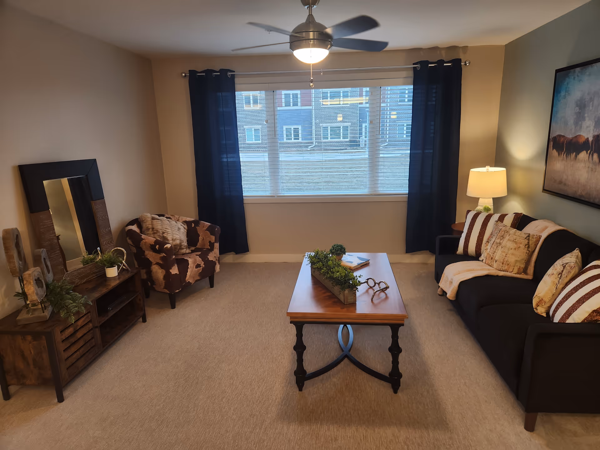 Well-lit living room with a sofa, armchair, coffee table, large window with dark curtains, and a ceiling fan.