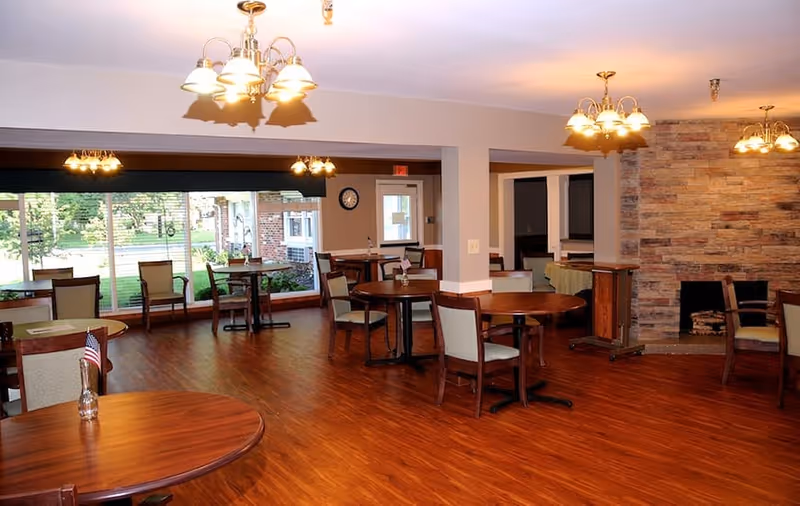 A spacious dining room with multiple round wooden tables and chairs arranged on a polished wooden floor. The room features large windows letting in natural light and a stone fireplace on one wall. Several ceiling light fixtures illuminate the area.