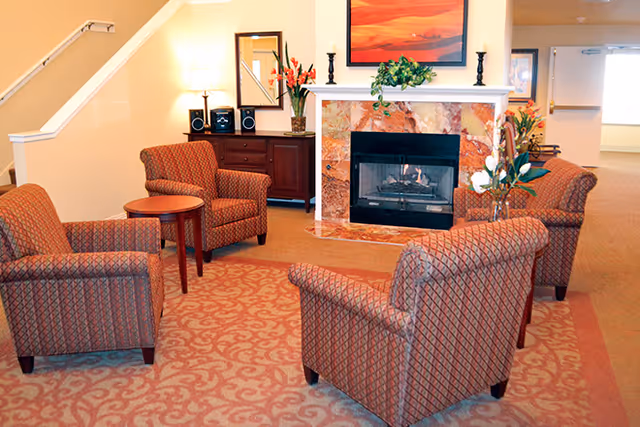 A cozy sitting area with four patterned armchairs arranged around a small table in front of a marble fireplace, with artwork, flowers, and a cabinet in the background.