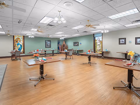 A spacious dining room with several tables set with napkins, plates, and flower centerpieces. The room has wooden flooring, green walls with framed pictures, and stained glass windows. Ceiling fans and fluorescent lights are mounted on the white tiled ceiling.