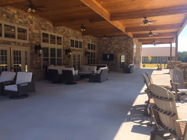 Covered outdoor patio area with stone walls and wooden ceiling beams. The space is furnished with cushioned wicker chairs and sofas arranged in seating groups, ceiling fans, and Adirondack chairs in the sunlight on the right side. Large windows and doors line the wall, and a TV is mounted on the stone wall.