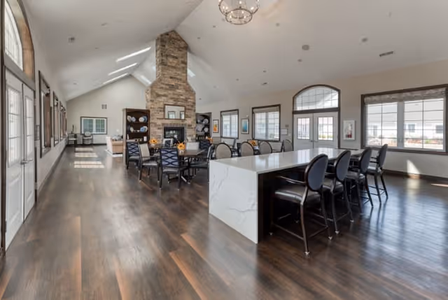 Spacious senior living common area with dark wood flooring, a large white marble island with black chairs, multiple dining tables with chairs, a stone fireplace, and large windows letting in natural light.