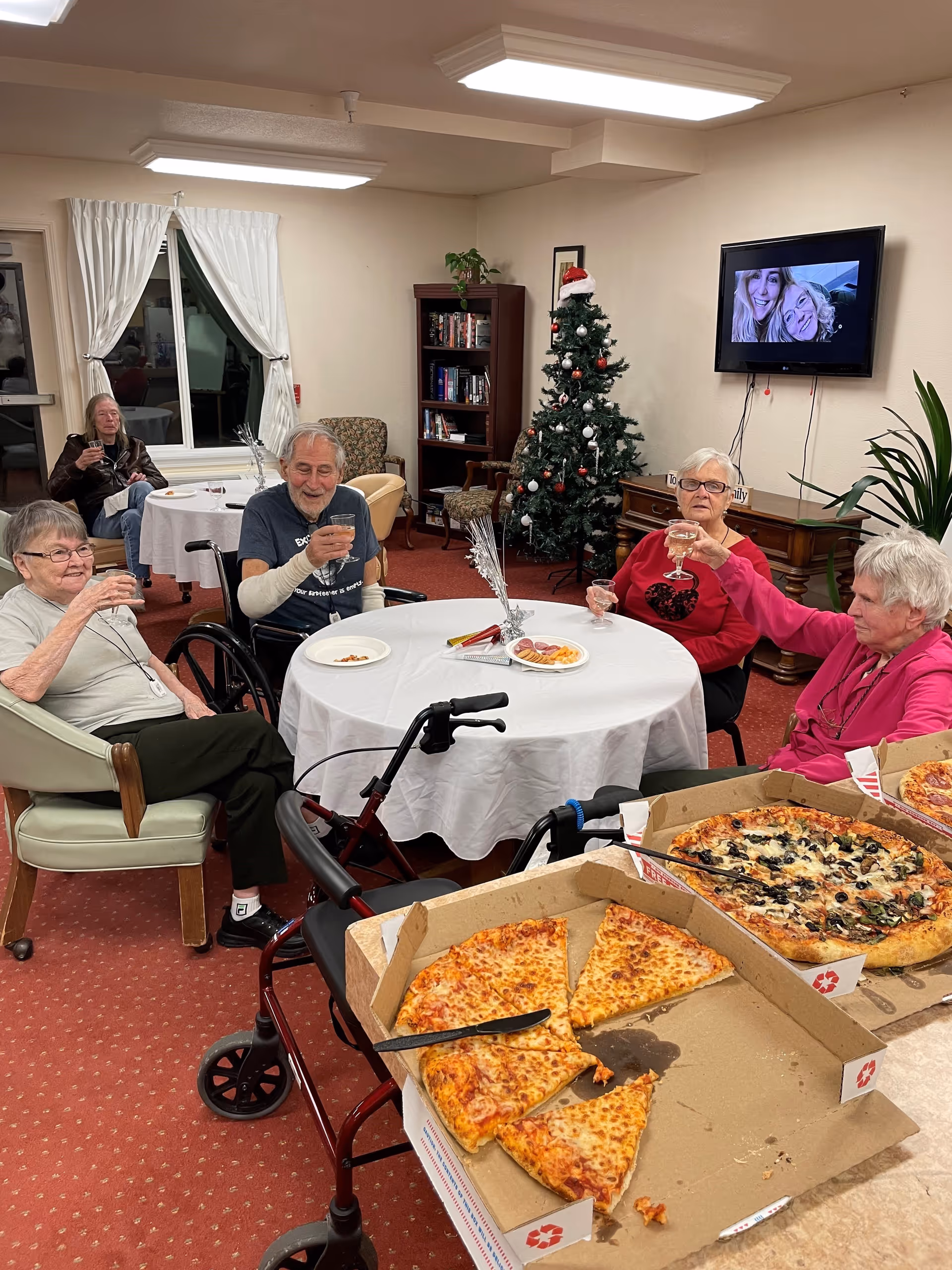 A group of elderly people sitting around a table in a senior living facility, enjoying drinks and pizza. The room is decorated with a Christmas tree and a TV mounted on the wall showing a photo of two smiling women. There are bookshelves and chairs in the background, and the atmosphere appears warm and festive.