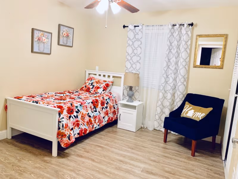 A cozy bedroom with a white wooden bed frame and a floral bedspread in shades of red, pink, and orange. Next to the bed is a white nightstand with a gray lamp. A navy blue armchair with a decorative pillow that says 'Home' is placed near a window with white and gray patterned curtains. The walls are light beige with two floral paintings and a gold-framed mirror. The floor is light wood, and a ceiling fan with lights is visible above.