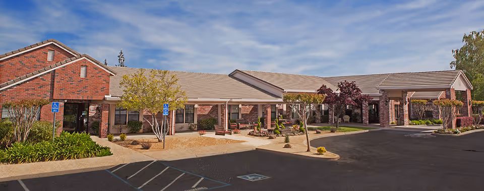 Front exterior of a single-story brick senior living facility with a covered entrance, landscaped courtyard, and parking area.
