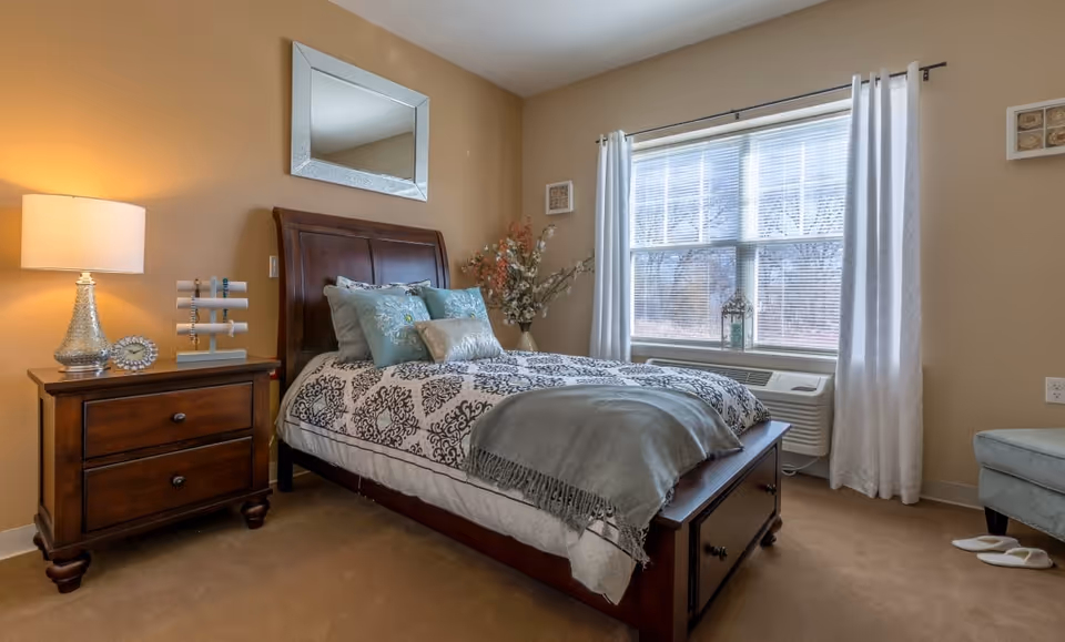 A cozy bedroom with a wooden bed frame and a patterned bedspread, decorated with multiple pillows. A wooden nightstand with a lamp and decorative items is beside the bed. A large window with white curtains lets in natural light, and a cushioned chair with slippers nearby is visible in the corner.