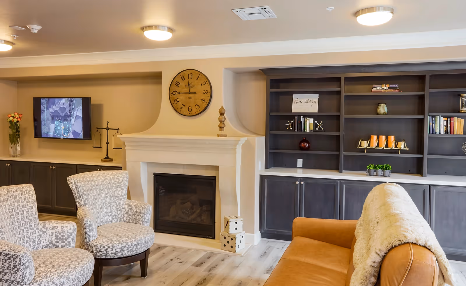 A cozy living room area in The Chalet featuring a tan leather sofa with a cream-colored throw blanket, two patterned armchairs, a white fireplace with a large round clock above it, built-in dark wood shelves with books and decorative items, and a flat-screen TV mounted on the wall.