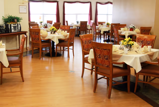 A dining room with several round tables covered with white tablecloths, each set with plates, cups, napkins, and floral centerpieces. Wooden chairs with patterned cushions surround the tables. Large windows with maroon curtains allow natural light to fill the room.