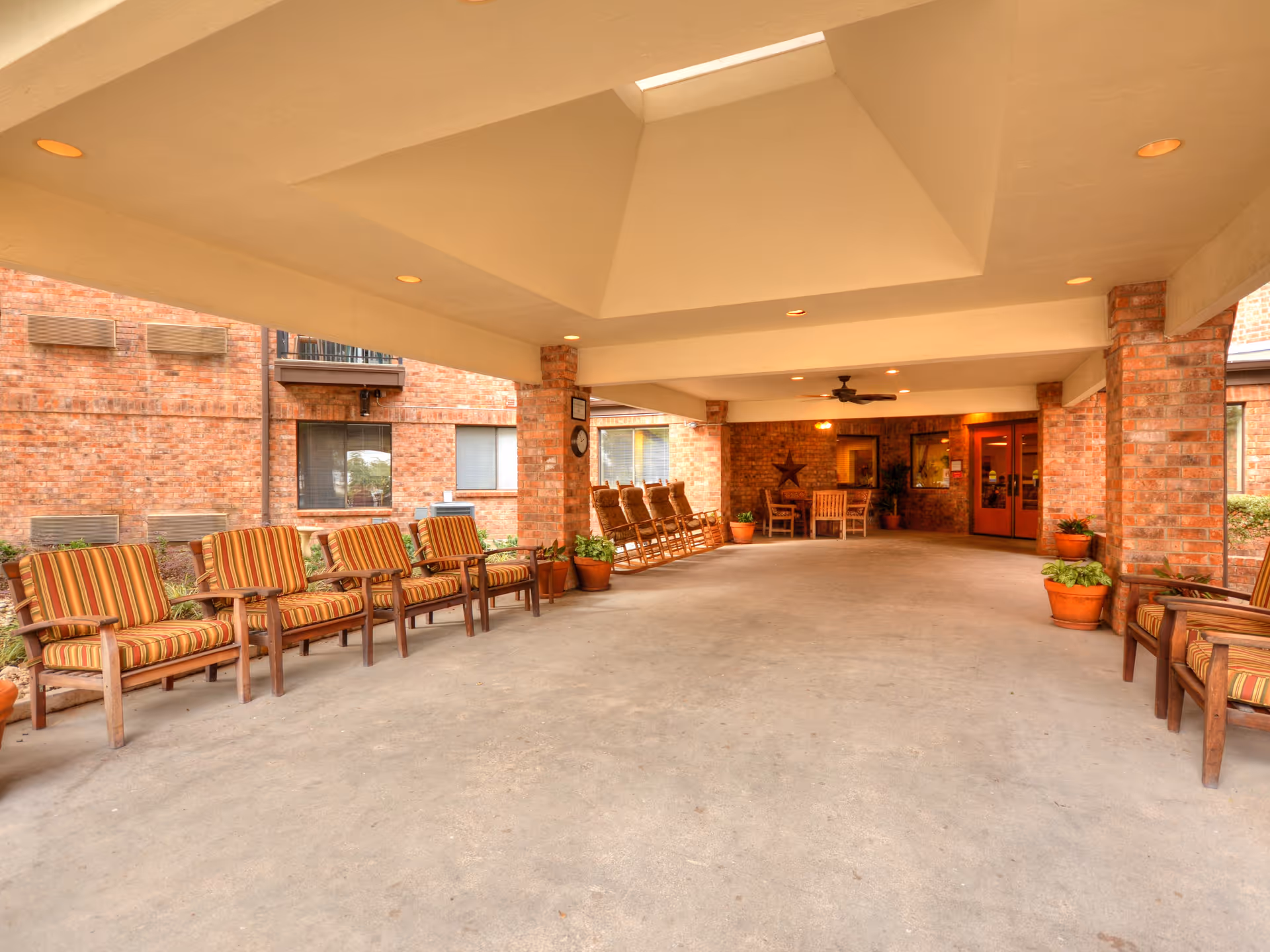 Covered outdoor seating area with striped cushioned wooden chairs lined up along brick walls, potted plants, and a table with chairs in the background near double doors.