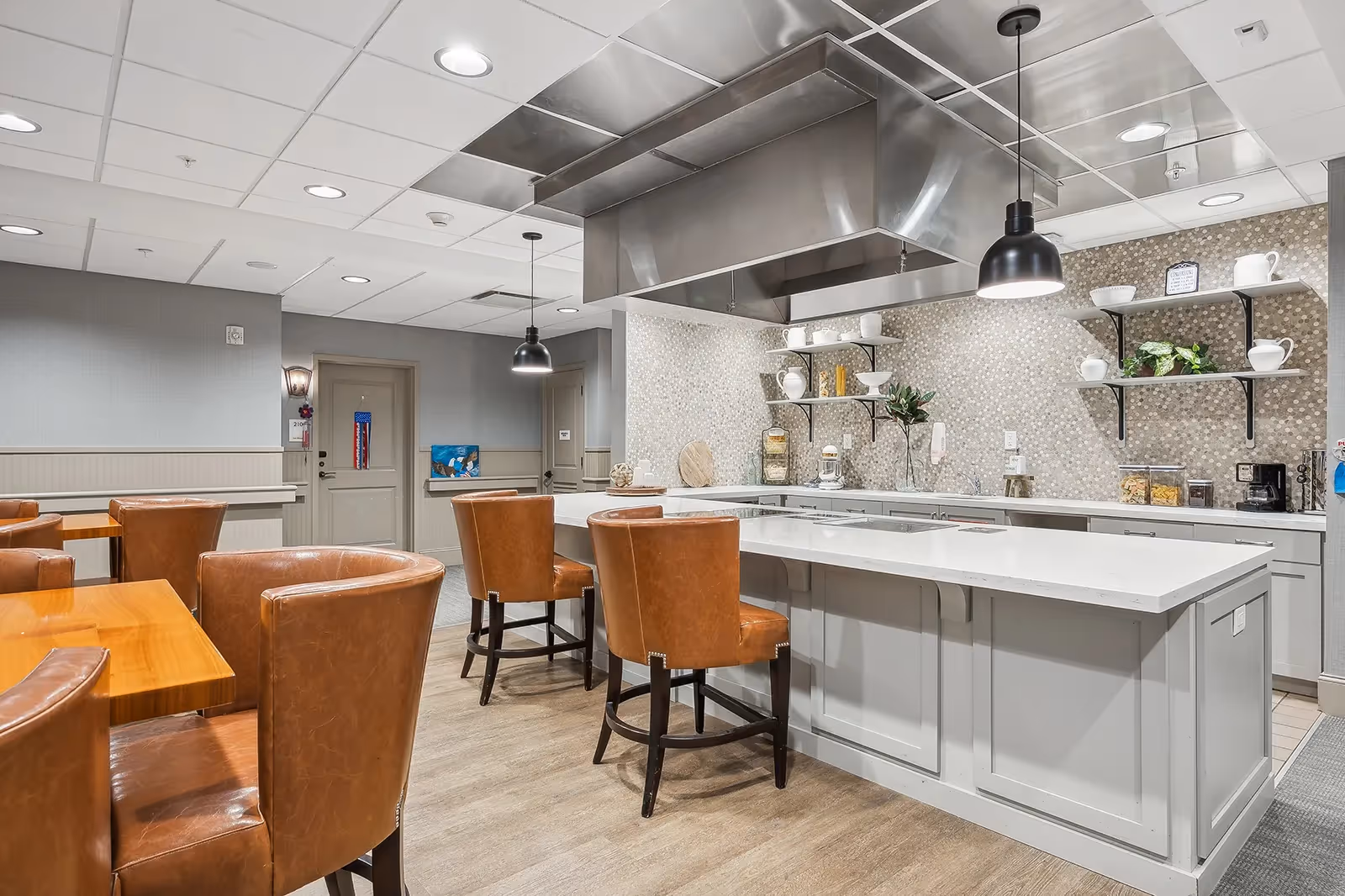 A modern kitchen and dining area with a large white island countertop surrounded by brown leather chairs. The kitchen features a stainless steel range hood, open shelves with white dishes and plants, and a mosaic tile backsplash. The dining area has wooden tables and matching brown leather chairs, with neutral-colored walls and ceiling lights.