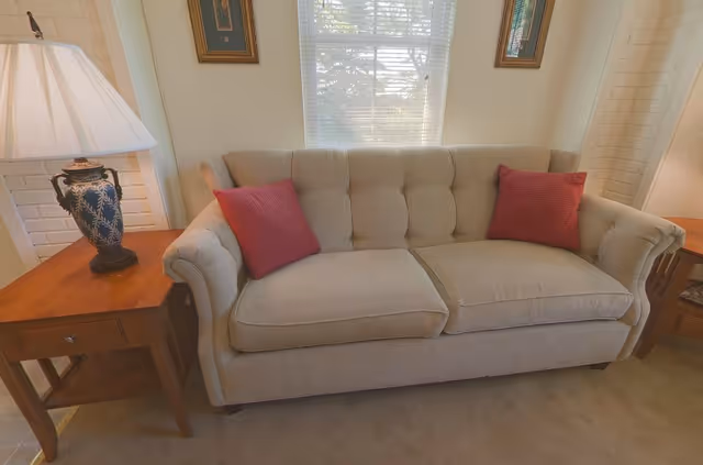 Beige tufted sofa with two red pillows flanked by wooden side tables and a lamp in front of a sunlit window.