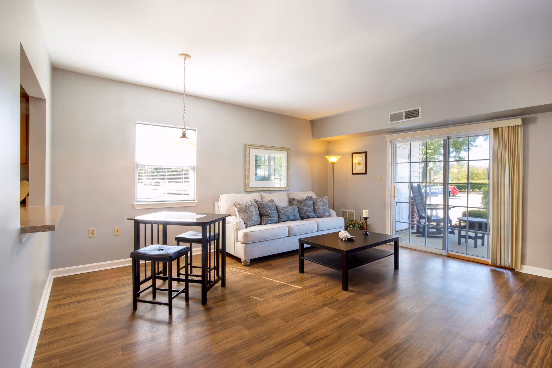 A bright living room with wooden flooring featuring a light gray sofa with multiple cushions, a black coffee table, a small dining table with two stools, a floor lamp, and a large sliding glass door leading to an outdoor patio area with chairs and a table.