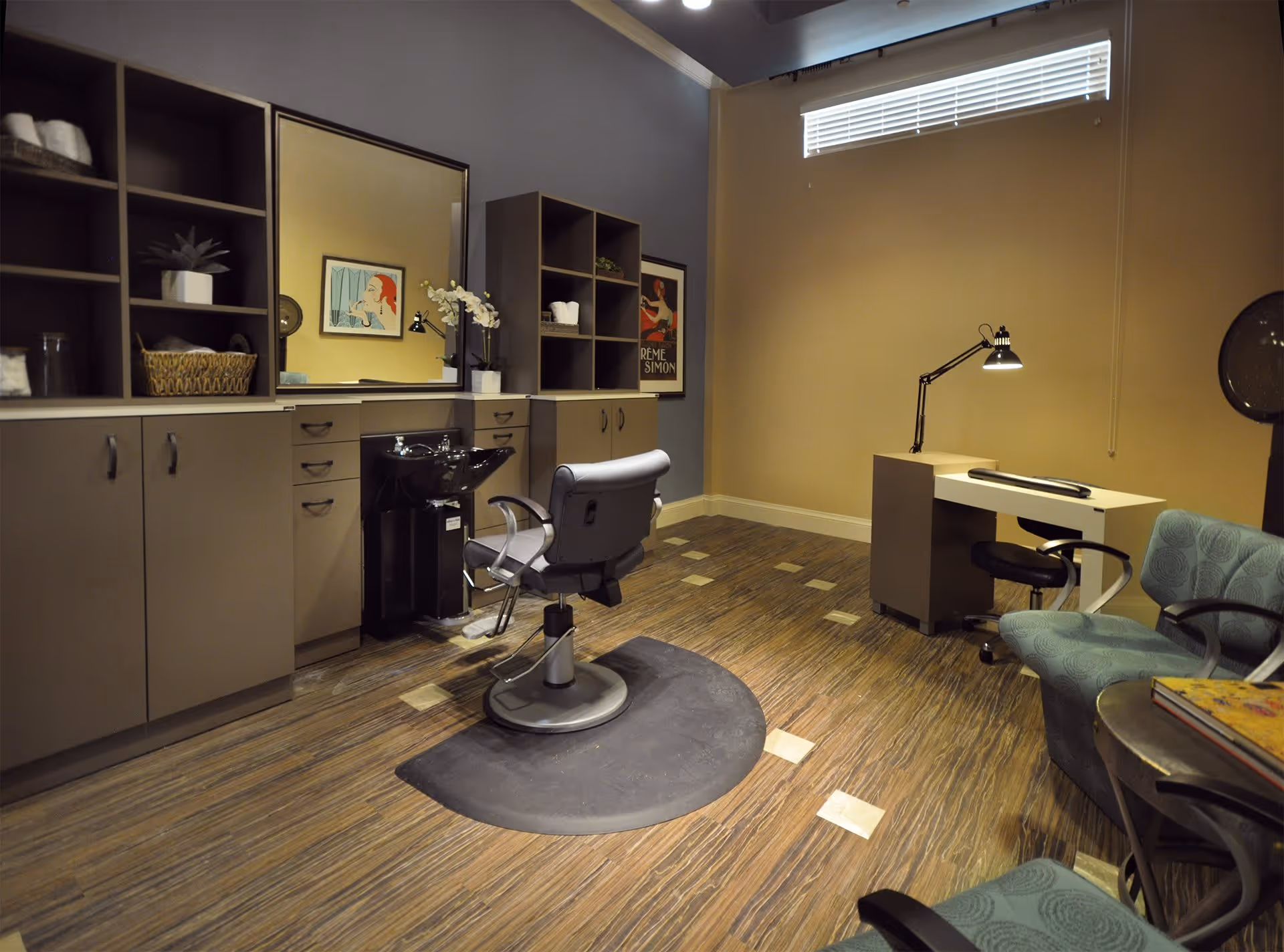 Interior view of a salon room with a styling chair in front of a large mirror and sink, cabinets with shelves holding towels and decor, a desk with a lamp and chair, and two upholstered chairs near a small round table.