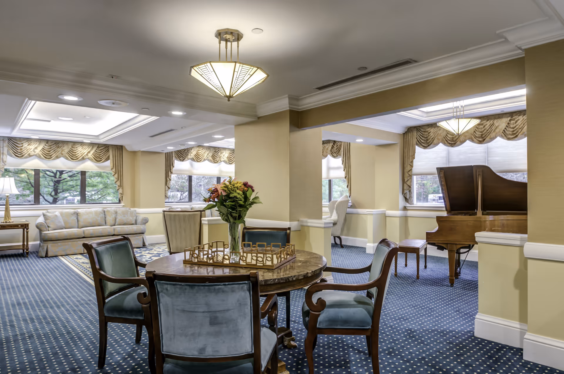 A spacious and elegant common area in a senior living facility featuring a round wooden table with four upholstered chairs, a vase with flowers on the table, a grand piano with a bench near the window, a sofa, armchairs, and large windows with decorative curtains letting in natural light.
