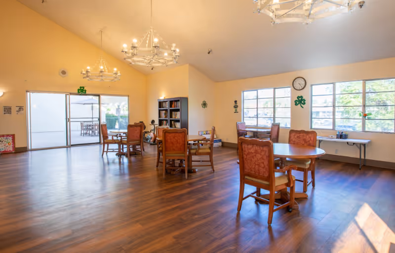 A spacious common area with wooden floors and several round tables surrounded by upholstered chairs. Large windows let in natural light, and there are two chandeliers hanging from the ceiling. A bookshelf and a clock are visible on the walls, and a sliding glass door leads to an outdoor patio with tables and umbrellas.