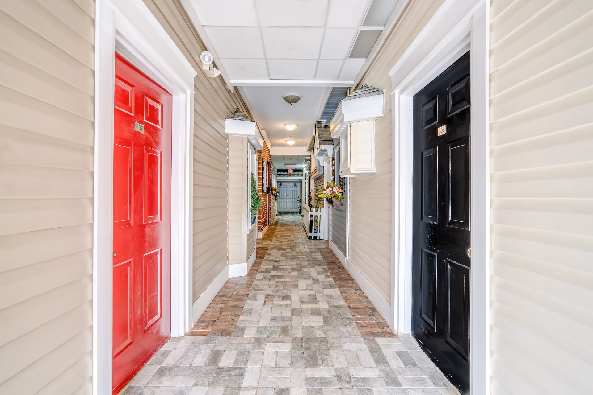 Indoor hallway with beige walls and tiled floor, featuring a red door labeled 307 on the left and a black door labeled 308 on the right. The hallway is decorated with small plants and flower boxes, and has ceiling lights and a white door at the far end.