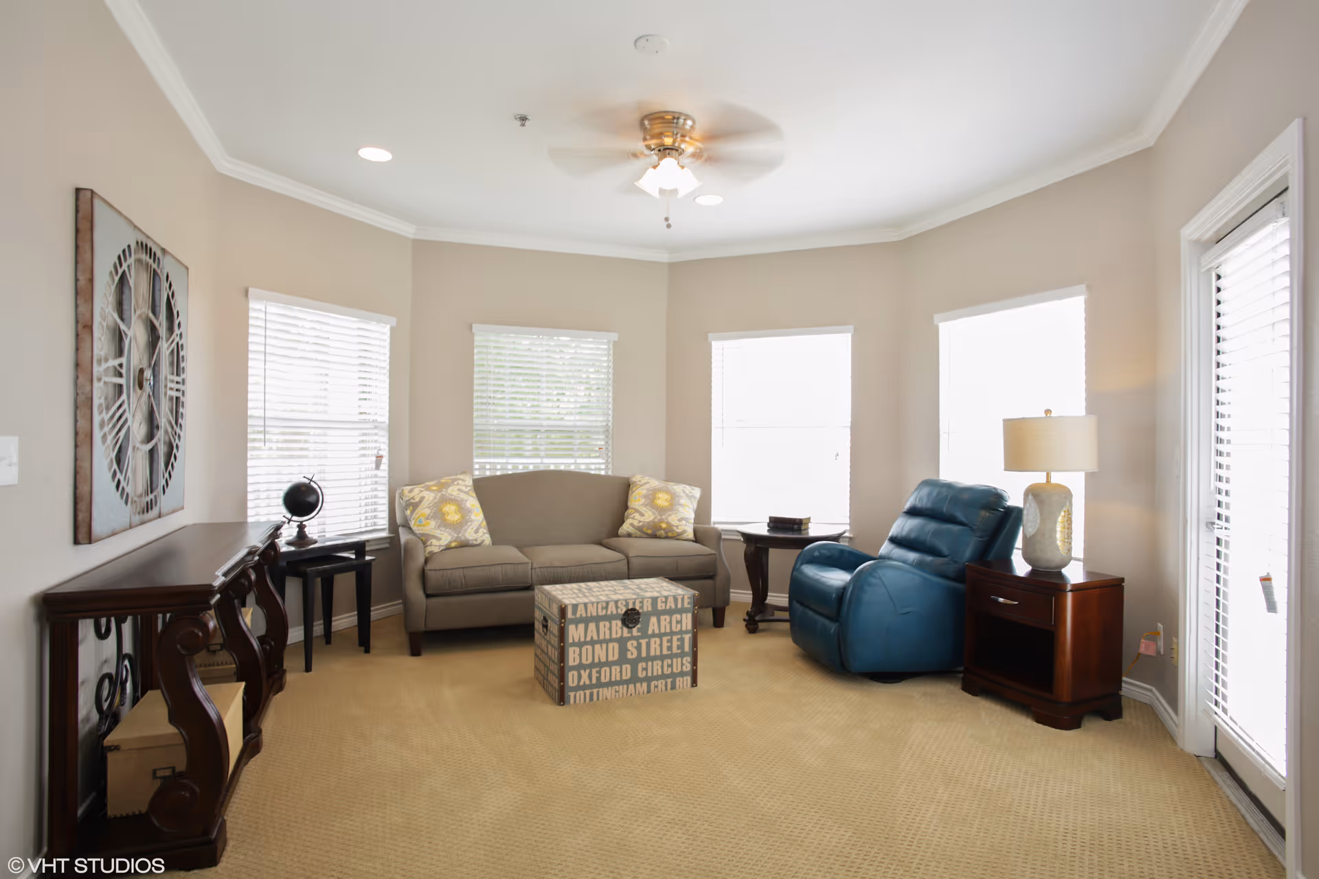 A bright living room with beige walls and carpet, featuring a gray sofa with two patterned pillows, a blue recliner chair, a wooden side table with a lamp, a small black side table with a globe, a decorative wall clock, and a rectangular ottoman with text printed on it. There are four windows with white blinds letting in natural light.