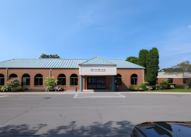 Front entrance of a one-story brick building with arched windows, a light blue metal roof and a covered drop-off area under a clear sky.