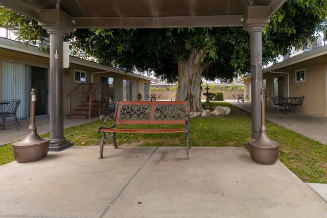 A decorative wooden bench sits under a covered patio facing a grassy courtyard with a large tree and single-story units on either side.