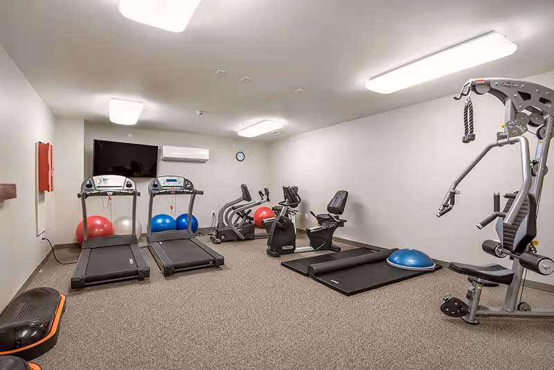 A fitness room with exercise equipment including two treadmills, two stationary bikes, a multi-function weight machine, exercise mats, balance balls, and a wall-mounted TV. The room has beige walls, carpeted floor, and bright overhead lighting.