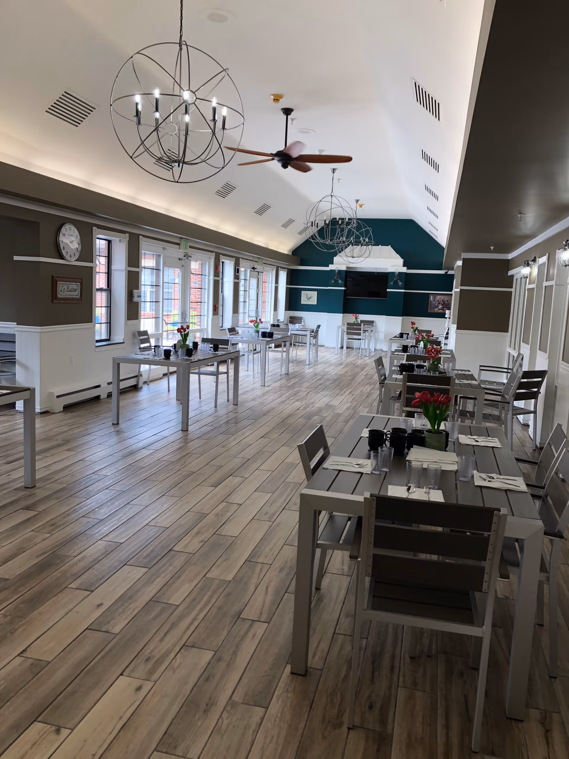 A spacious dining room with wooden flooring, several tables and chairs arranged neatly, each table set with napkins, utensils, glasses, and small flower vases. The room has large windows along one side, modern spherical chandeliers hanging from the ceiling, and a ceiling fan. The far wall is painted teal with a mounted TV and decorative artwork.