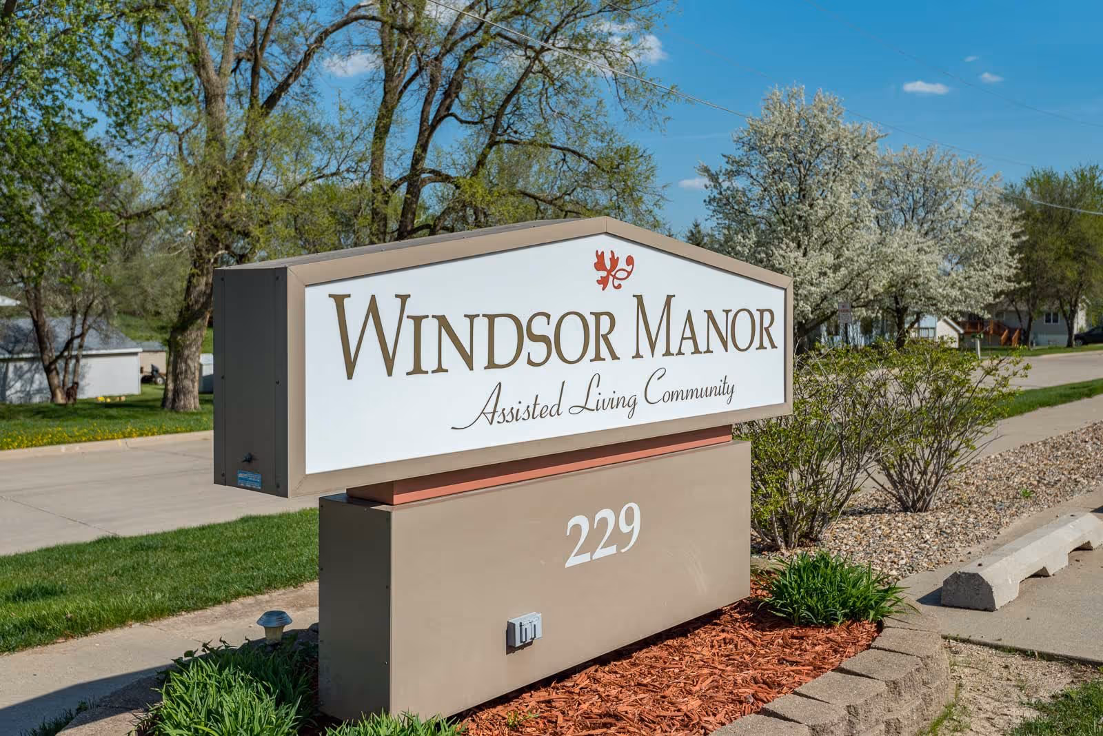 Outdoor sign for Windsor Manor Assisted Living Community with the address number 229, surrounded by greenery and trees under a clear blue sky.