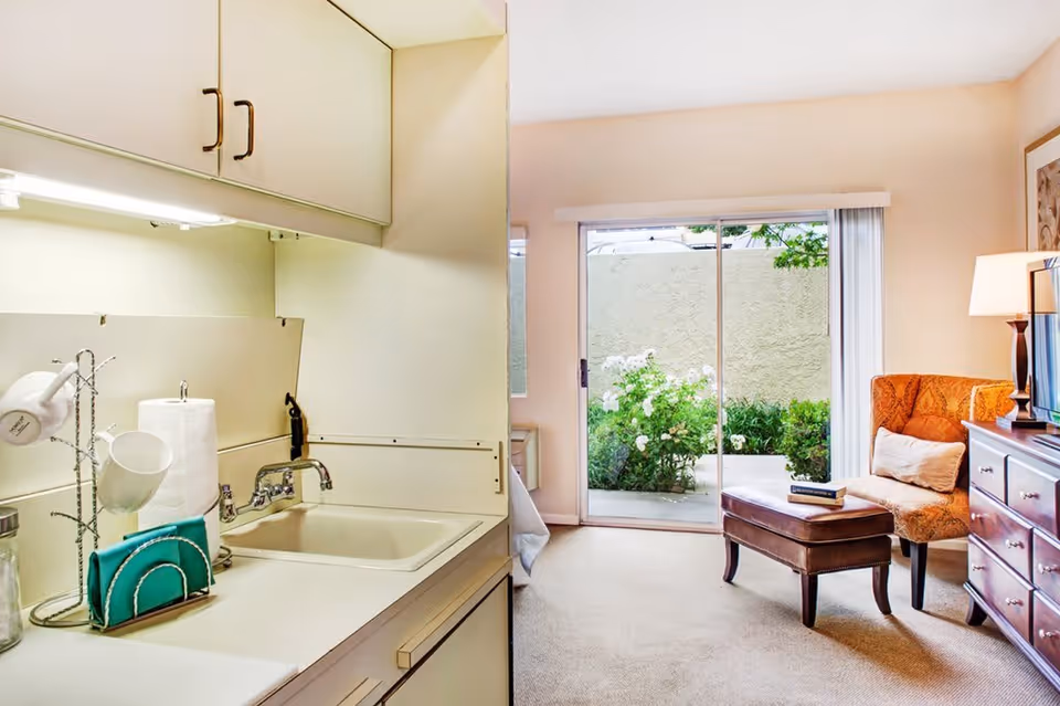 A small kitchenette area with a sink, paper towel holder, and hanging cups on the left side, leading into a cozy living space with an orange patterned armchair, a brown ottoman with books on it, a wooden dresser with a lamp, and a sliding glass door opening to a small outdoor garden area with white flowers and greenery.