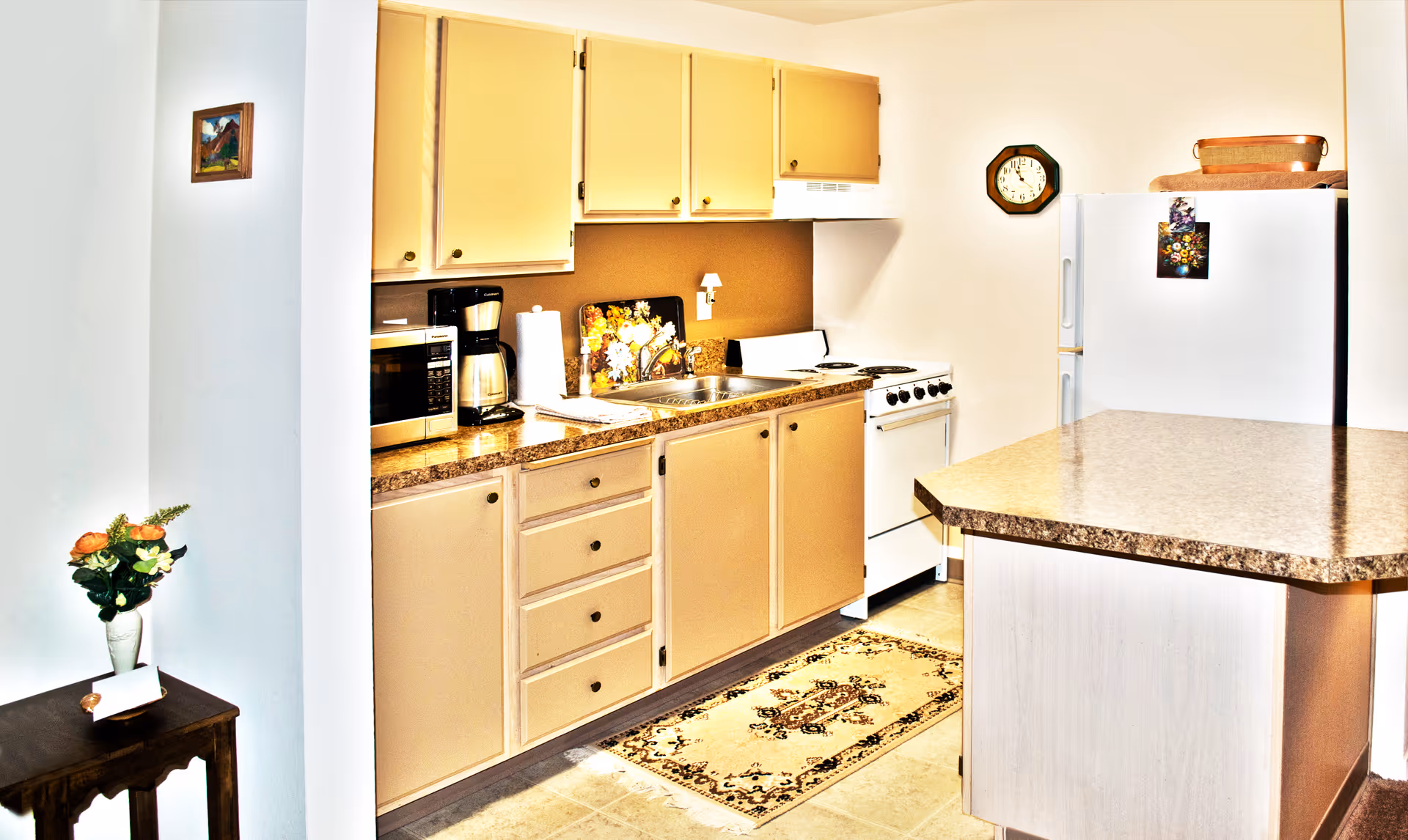 A bright kitchen with beige cabinets and drawers, a countertop with a coffee maker, microwave, and paper towel holder. There is a white stove and refrigerator, a wall clock, and a small decorative rug on the floor. A small table with a vase of flowers and a framed picture on the wall are visible on the left side.