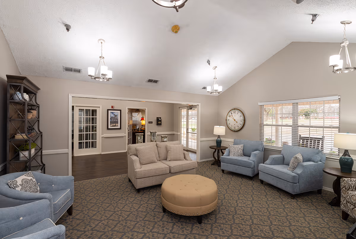 A cozy living room area with beige and blue upholstered chairs and sofas arranged around a round beige ottoman. The room has a patterned carpet, light gray walls, and large windows with white blinds letting in natural light. There are two side tables with lamps, a large wall clock, and a wooden shelving unit with decorative items. The ceiling is vaulted with modern light fixtures.