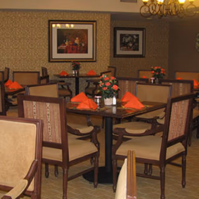 Dining room with multiple wooden tables and cushioned chairs. Each table is set with orange folded napkins and a small flower arrangement in the center. The walls have patterned wallpaper and framed artwork, and a chandelier hangs from the ceiling.