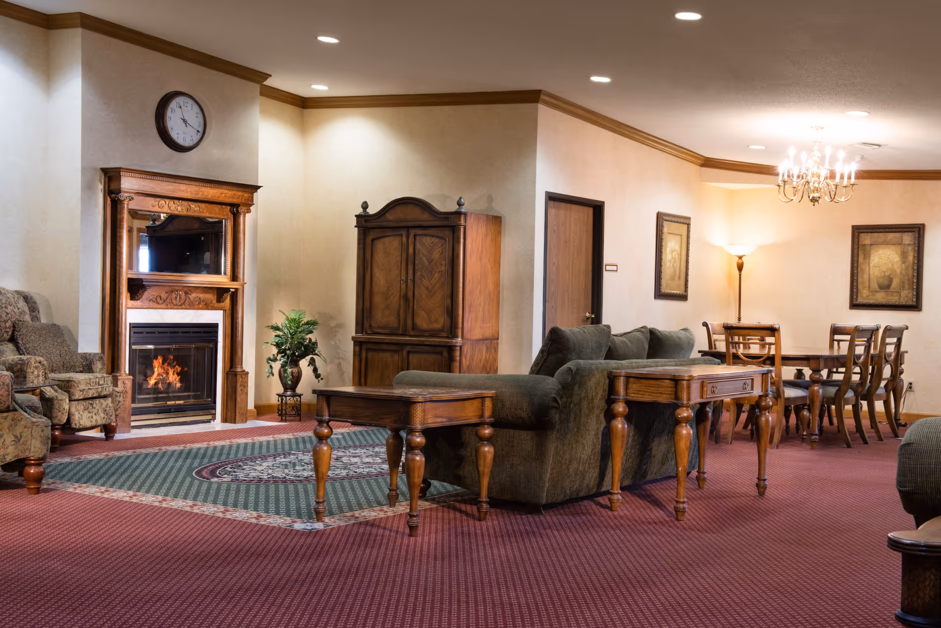 A cozy senior living common area with a lit fireplace surrounded by a wooden mantel and mirror. The room features a green sofa, wooden side tables, armchairs, a dining table with chairs, framed artwork on the walls, a floor lamp, and a clock above the fireplace. The carpet is red with a green patterned rug in front of the fireplace.